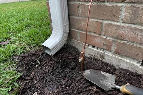 Bare copper conductor clamped to a ground rod at the base of an exterior wall, in mulch