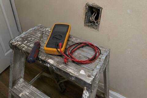 Multimeter resting on the top step of a ladder in front of an open wall box