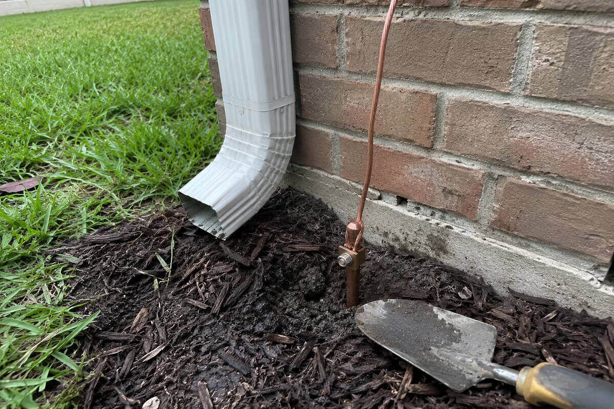 Bare copper conductor clamped to a ground rod at the base of an exterior wall, in mulch