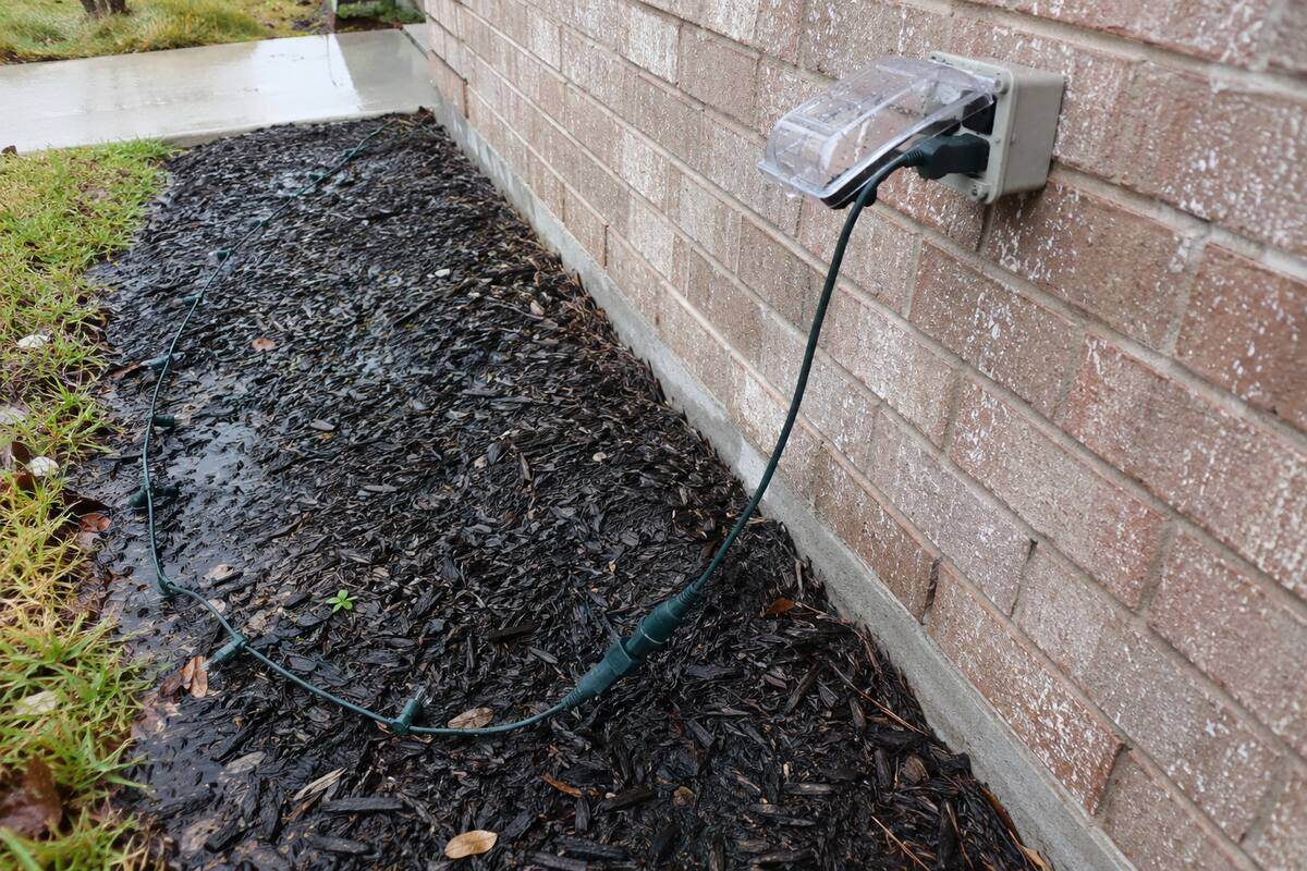 Exterior receptacle on a brick wall with its in-use cover propped open by a light string plug, water beading on the cord